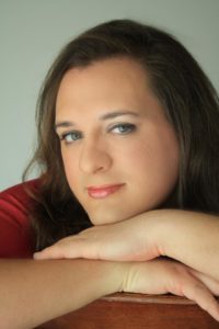 Portrait of a person with long dark hair and light makeup, resting their chin on folded arms and looking toward the camera.
