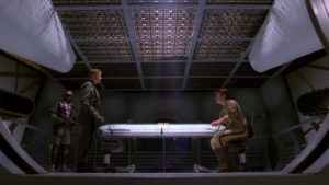 Two men face each other across a bright table in a stark metal room.