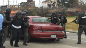 SWAT officers detain a person beside a red sedan during a vehicle assault demonstration.